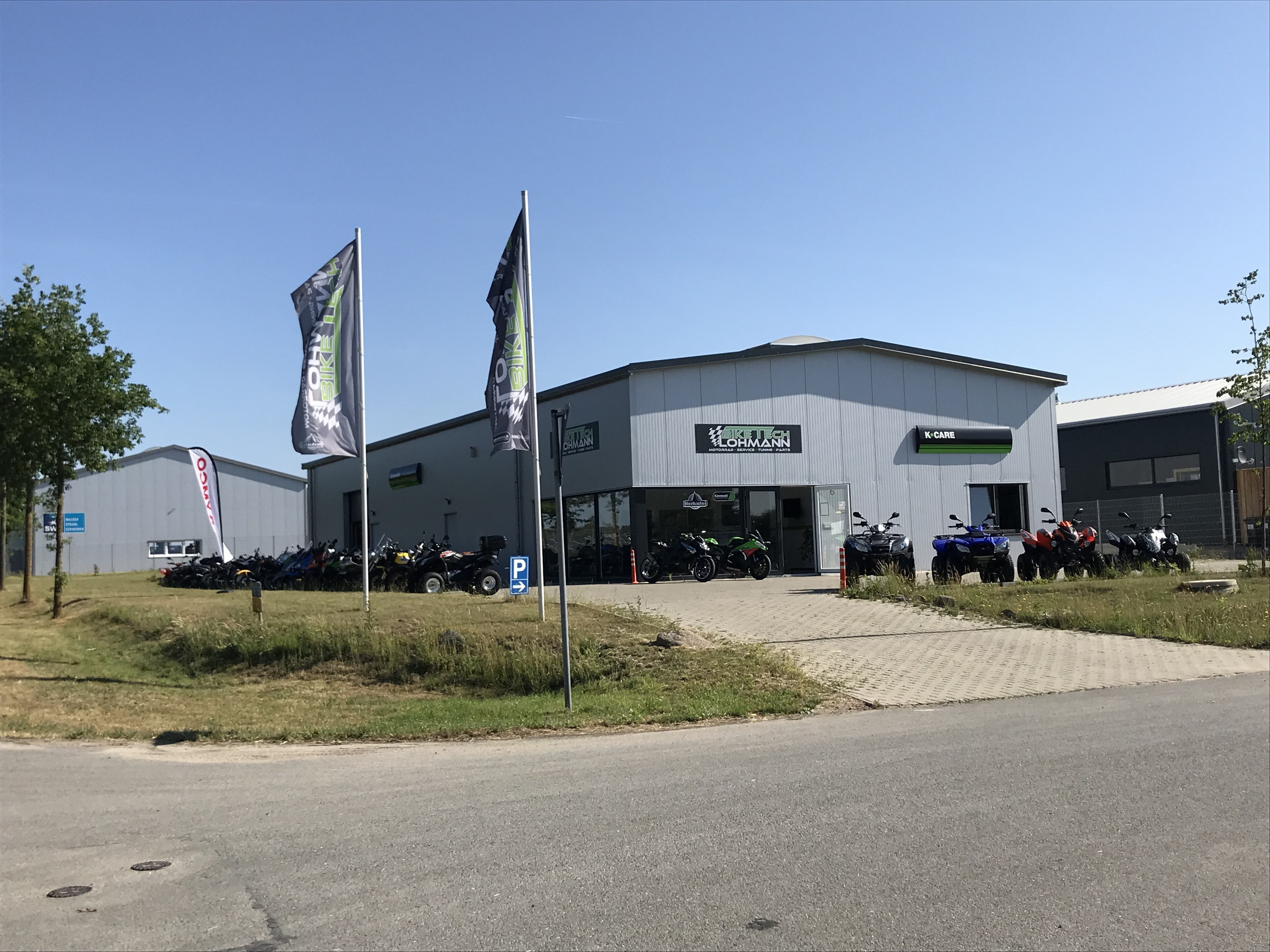 BikeTech Lohmann Außenansicht – Werkstatthalle mit Auffahrt, Fahrzeugen auf dem Hof und blauem Himmel